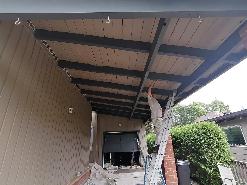 Professional Ottawa painter on a tall extension ladder painting the complex underside and exposed beams of a large covered carport ceiling. NCR Pro-Painting team uses drop cloths and safety equipment for high-ceiling residential projects.