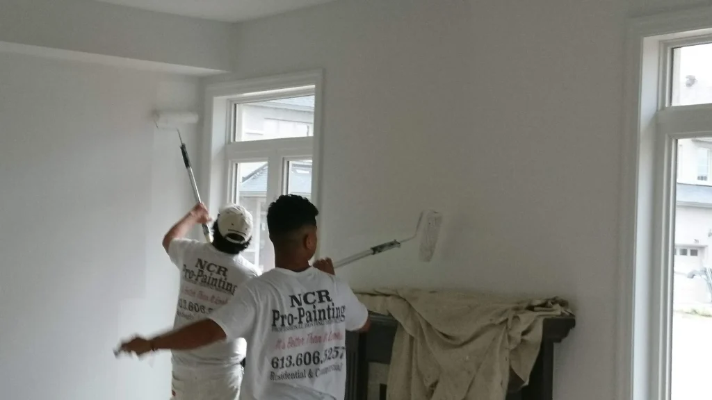 Two professional painters from NCR Pro-Painting in uniform applying white wall paint using extension poles, with full floor protection and paint cans in a modern Ottawa living room.